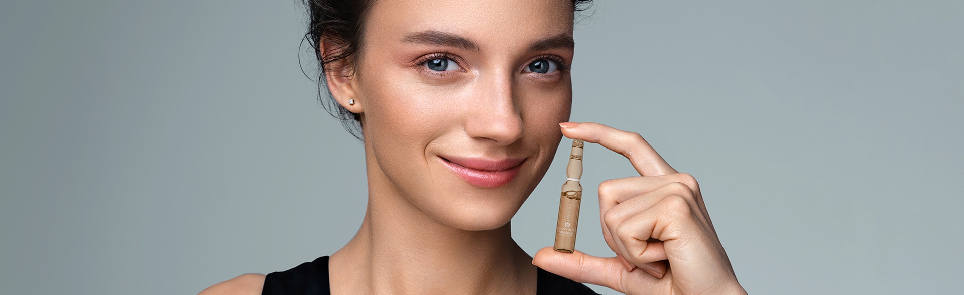 Frau mit zurückgebundenen Haaren im schwarzen Top hält lächelnd eine Glasampulle.